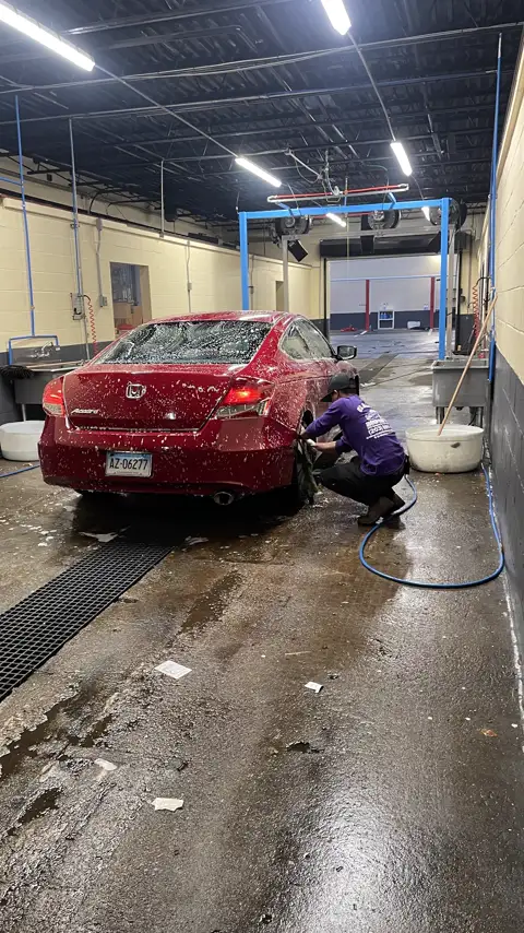 A red Honda Accord covered in soap in a wash bay while a worker scrubs the rear wheel