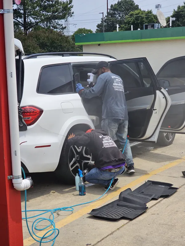 Two workers cleaning a white Mercedes SUV by hand: one wipes the inside of a rear door, one scrubs a wheel, with the floor mats laid out to dry