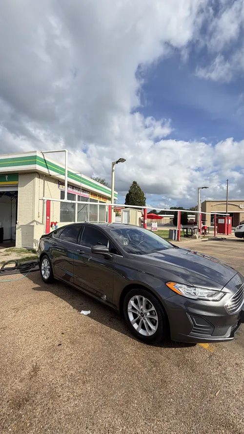 A gray Ford Fusion on the lot in front of the green-striped building and red posts